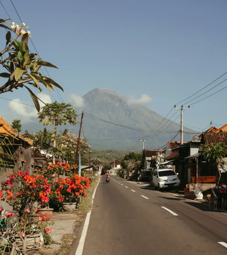 Vista del volcán Agung desde una carretera en Amed, Bali, rodeada de casas tradicionales, plantas con flores y tráfico local.