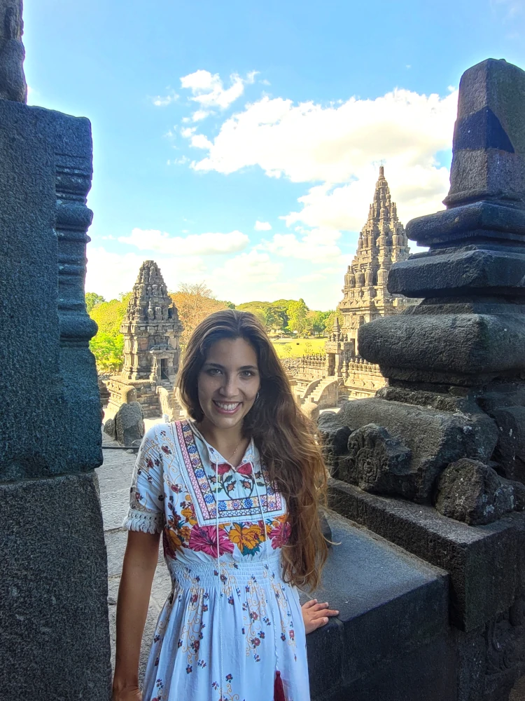 En el templo Prambanan, de pie entre estructuras de piedra, con varios santuarios hinduistas visibles al fondo bajo un cielo despejado.