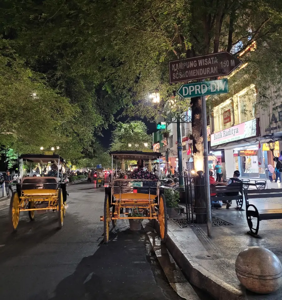 Malioboro Street de noche con becaks tradicionales circulando entre tiendas iluminadas, peatones y ambiente animado de fin de semana.